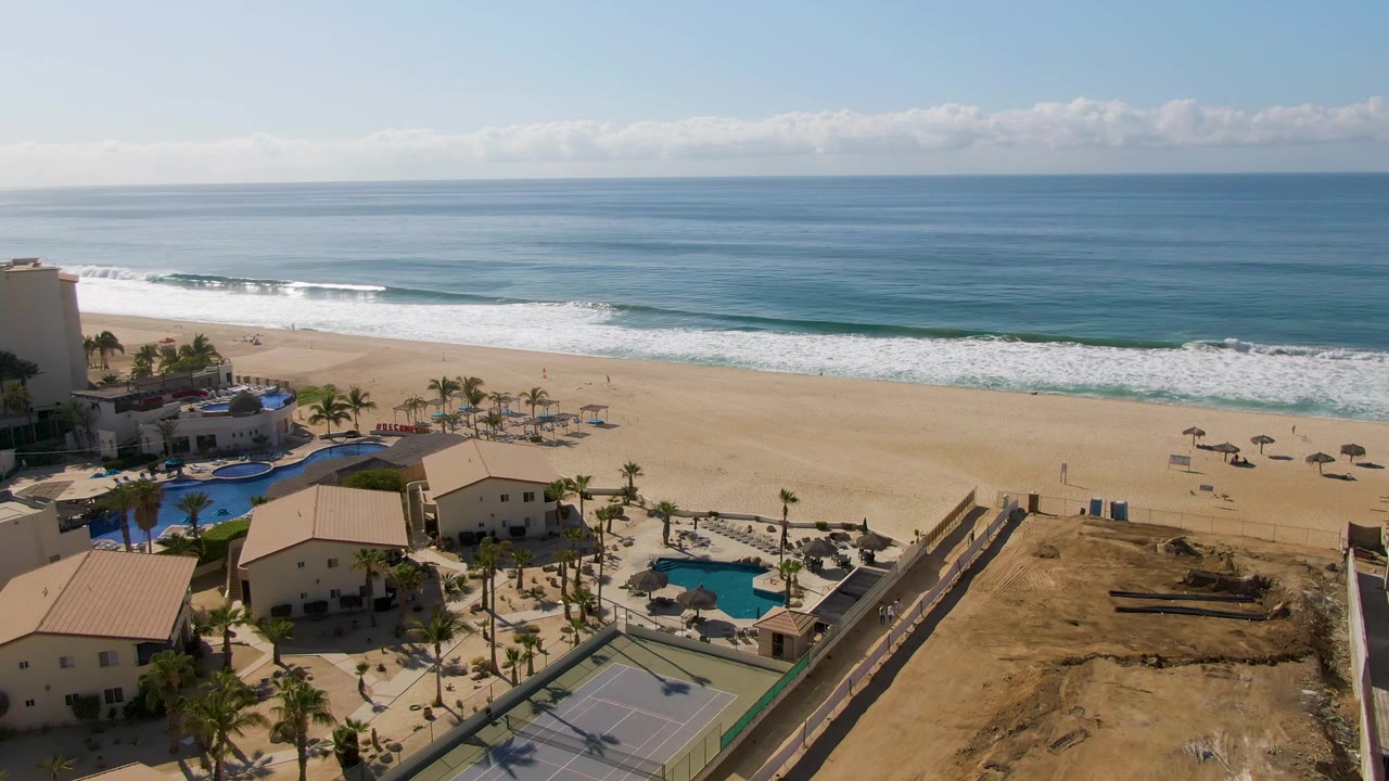 El Tule Beach appears from above with a level ocean horizon, broad sandy shoreline, palm-lined resort grounds, a visible pool area, tennis court, and no visible people.