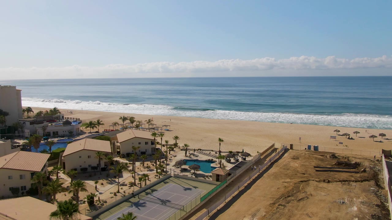 Opening aerial frame over El Tule Beach in Los Cabos, Mexico, with resort buildings, pools, tennis courts, palms, sandy beach, ocean waves, and no visible people.