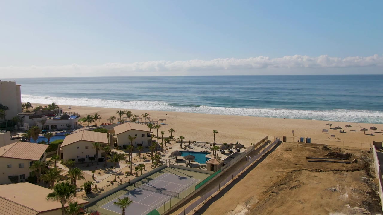 Aerial view at 0:01 over El Tule Beach with the tennis courts more centered below, pools and palms beside resort buildings, surf on the sand, and no visible people.