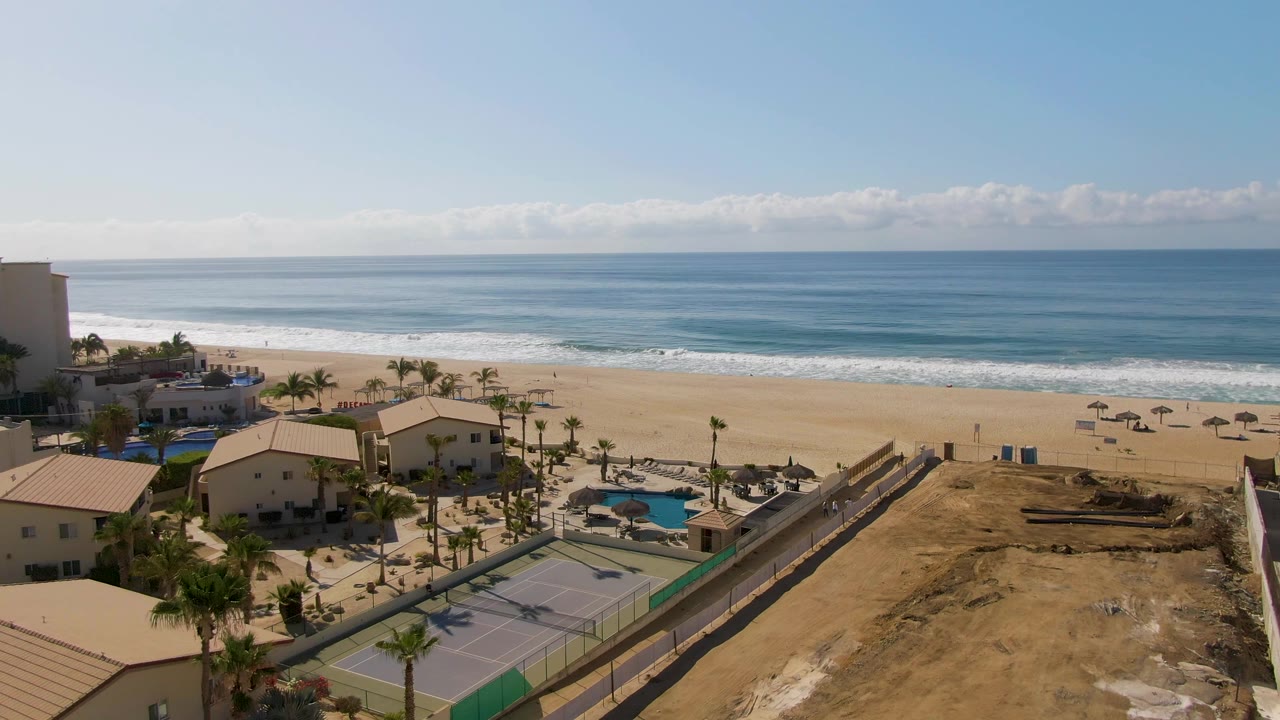 At 0:02, the El Tule Beach aerial shows resort roofs, palm trees, pools, tennis courts, beach umbrellas, blue ocean, and no visible people under clear sun.