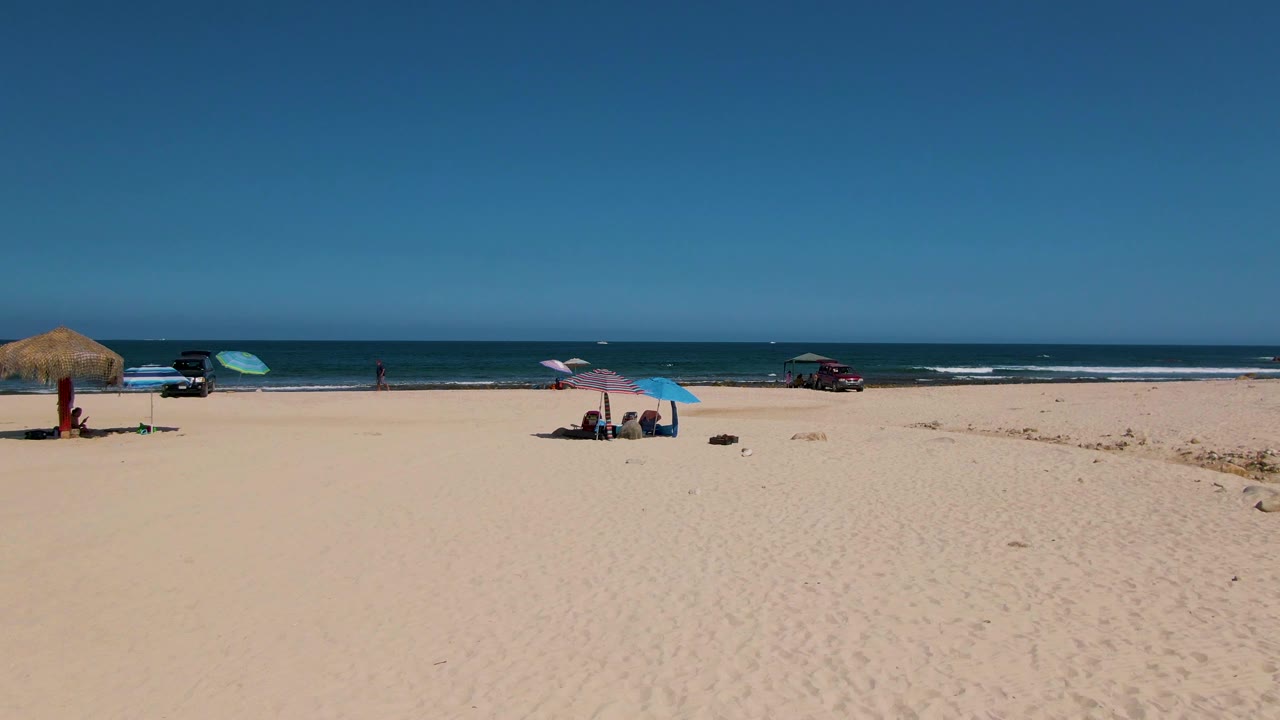 Wide aerial frame of El Tule Beach in Los Cabos with pale sand, a thatched umbrella at left, colored umbrellas near the center, distant people, vehicles by the shoreline, blue ocean, and clear sky.