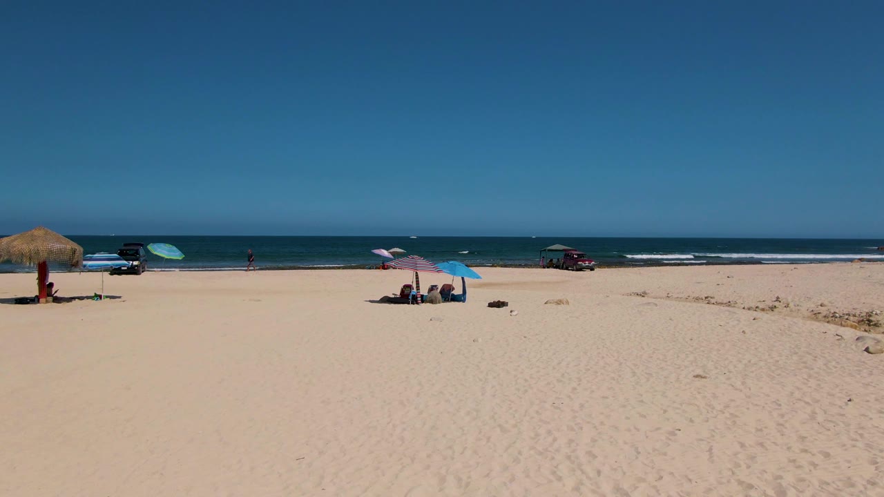 Aerial view over El Tule Beach in Los Cabos with a thatched shade closer to center-left, a blue umbrella near seated people, vehicles near the waterline, low waves, and a clear horizon.
