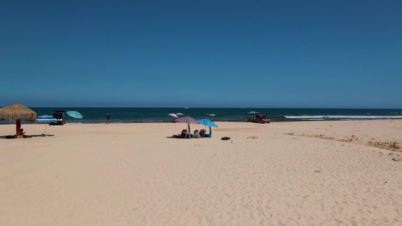 El Tule Beach aerial frame with broad empty sand in front, a thatched umbrella left of center, a blue umbrella sheltering people, a red vehicle near the right shoreline, and calm blue ocean beyond.