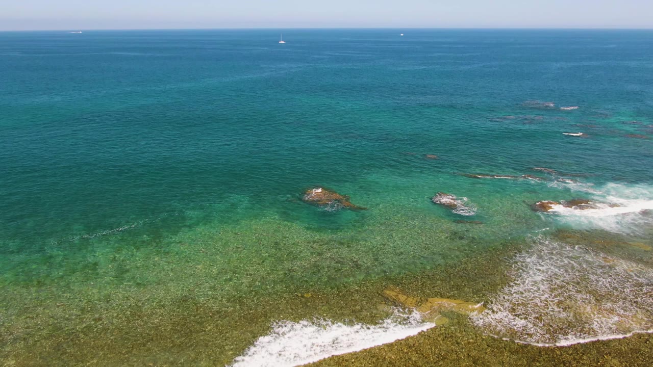 Aerial frame at El Tule Beach, Los Cabos showing turquoise water, shallow rock shelves, white surf near the lower edge, distant boats on the horizon, and no visible people.