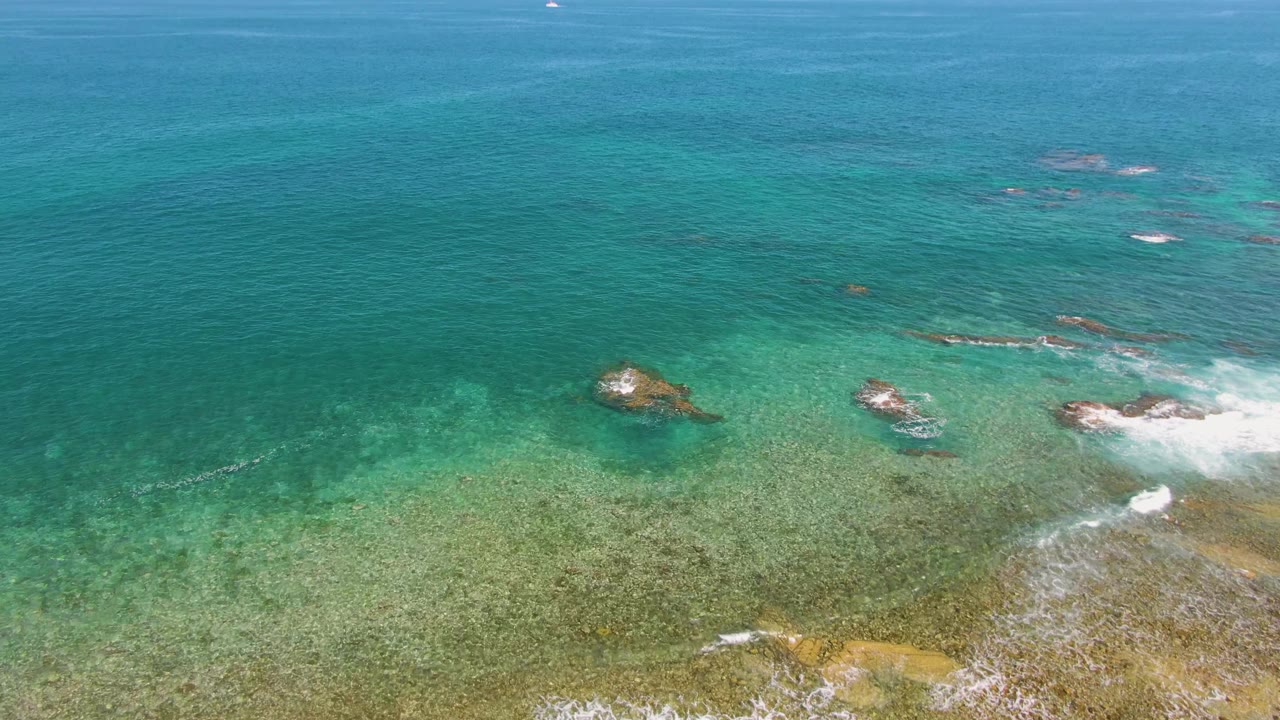 Aerial frame at El Tule Beach, Los Cabos showing shallow reef-colored water across the lower frame, scattered exposed rocks, narrow whitecaps near the right edge, and no visible people.