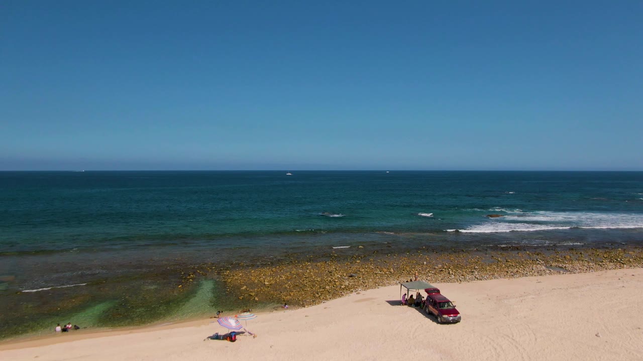 Aerial frame at El Tule Beach in Los Cabos showing pale sand with a red vehicle, umbrella, small visible people, rocky shallow water, and the blue ocean beyond.