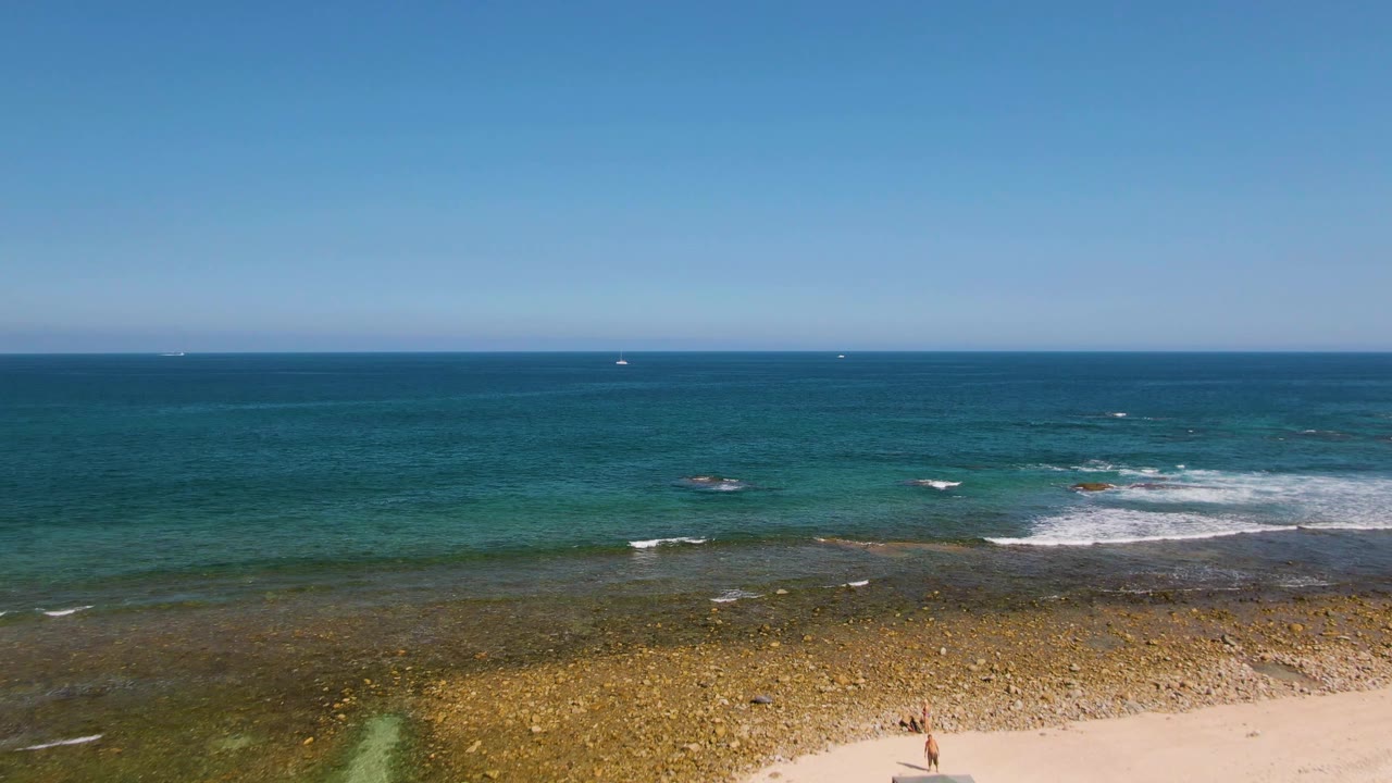 Aerial view of rocky shoreline, small strip of sand, turquoise water, white surf, and open ocean.