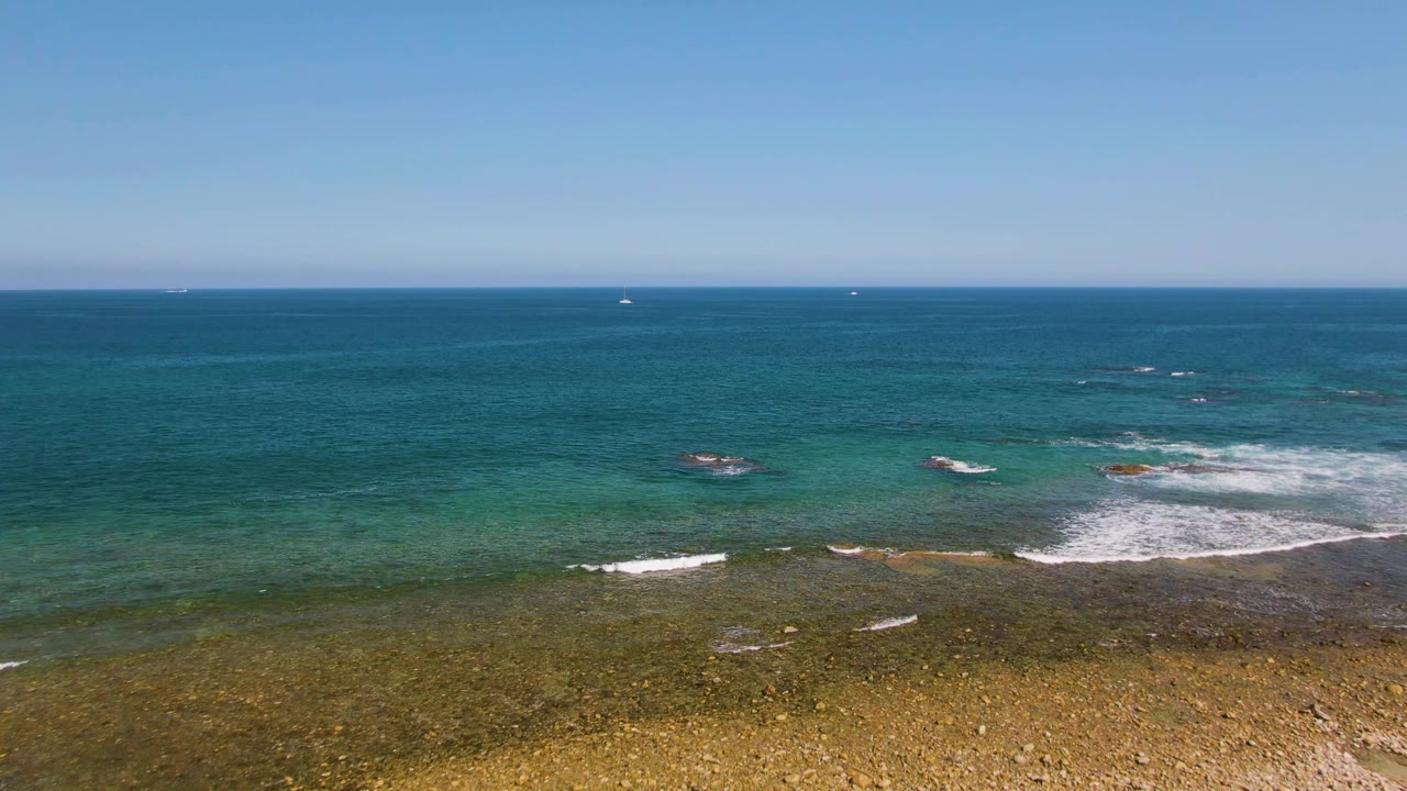 Aerial frame at El Tule Beach in Los Cabos centered on rocky shallows and turquoise water, with the sandy beach mostly out of frame and no visible people.