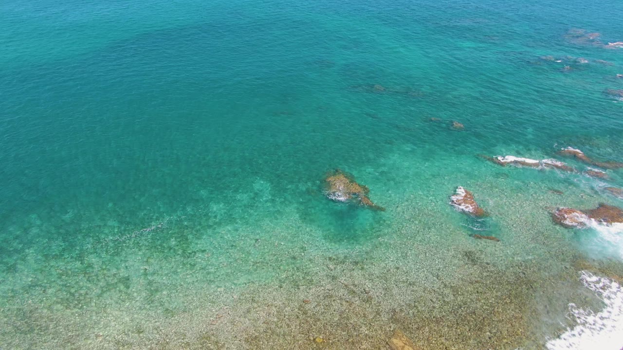 Aerial view at El Tule Beach in Los Cabos showing turquoise water over submerged rocks, with small foam streaks near the lower right and no visible people.