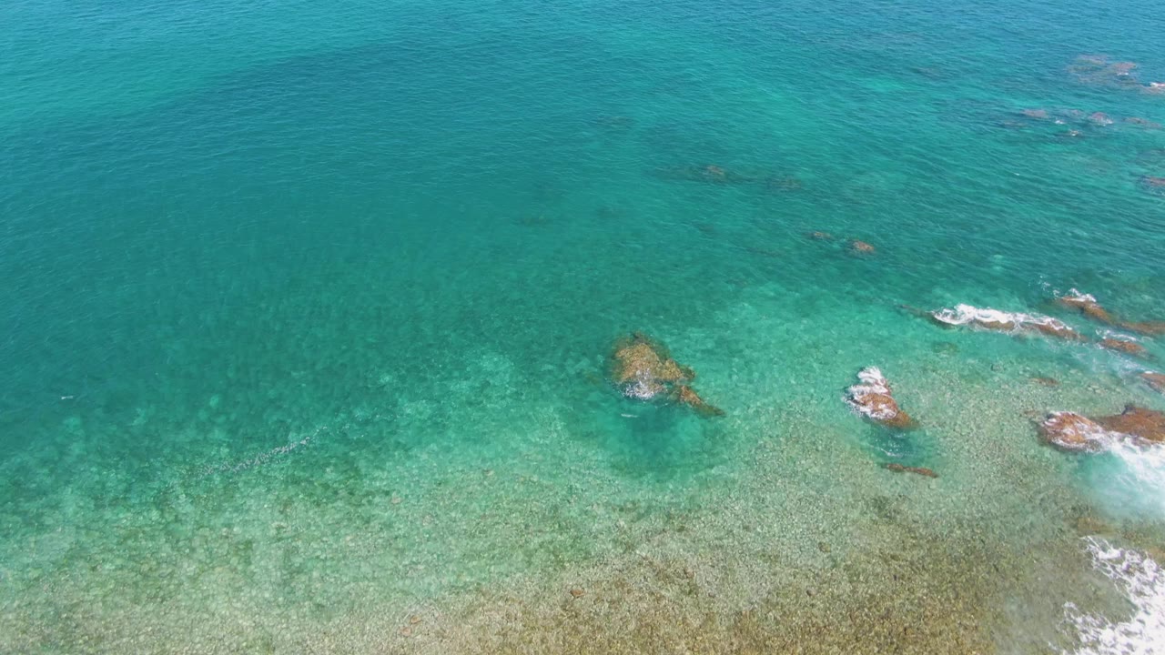 Aerial frame from El Tule Beach in Los Cabos with pale green shallows, darker turquoise water above, exposed rock shapes to the right, and no visible people.