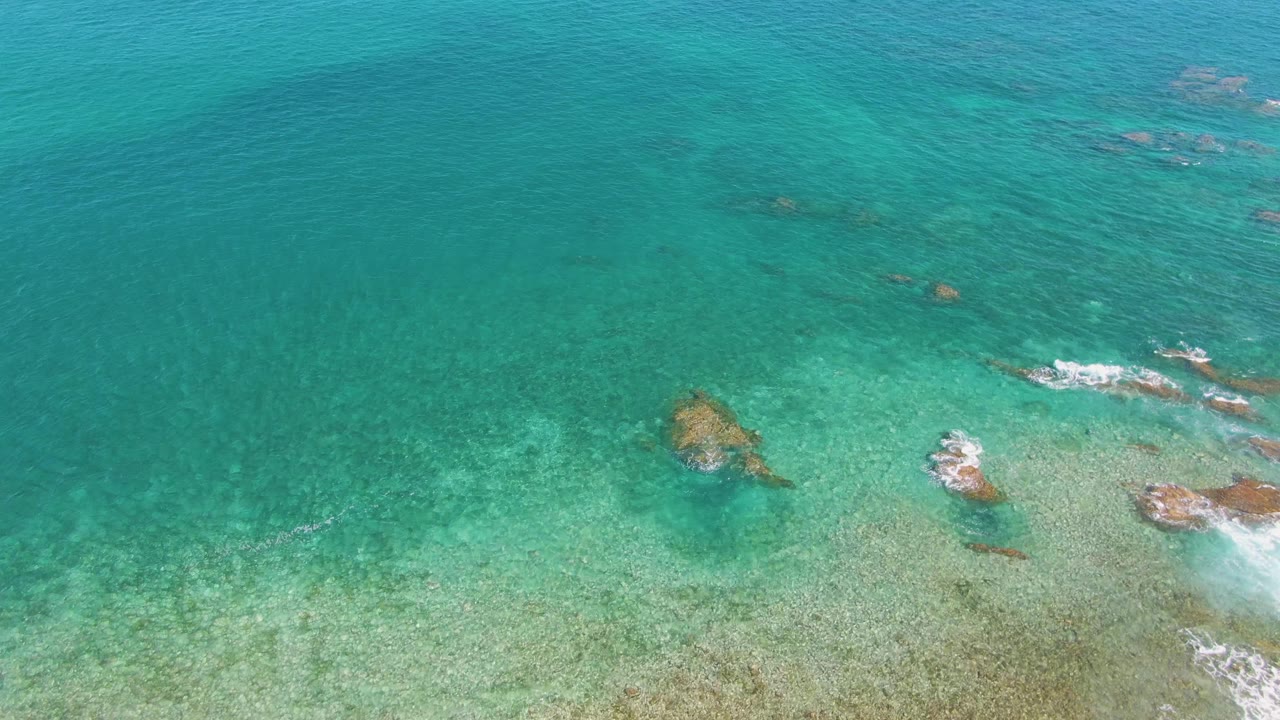 Overhead coastal water at El Tule Beach in Los Cabos shows clustered submerged rocks near the right side, a lighter sandy seabed below, and no visible people.