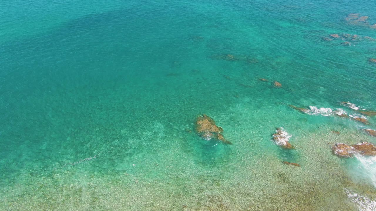 Turquoise ocean surface with a central submerged rock and foam near the bottom right.