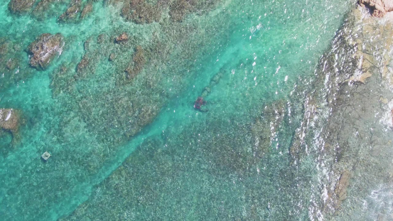 Overhead view at El Tule Beach Los Cabos showing clear turquoise shallows, dark reef patches, submerged rocks, and a small swimmer form near the center with no recognizable people.