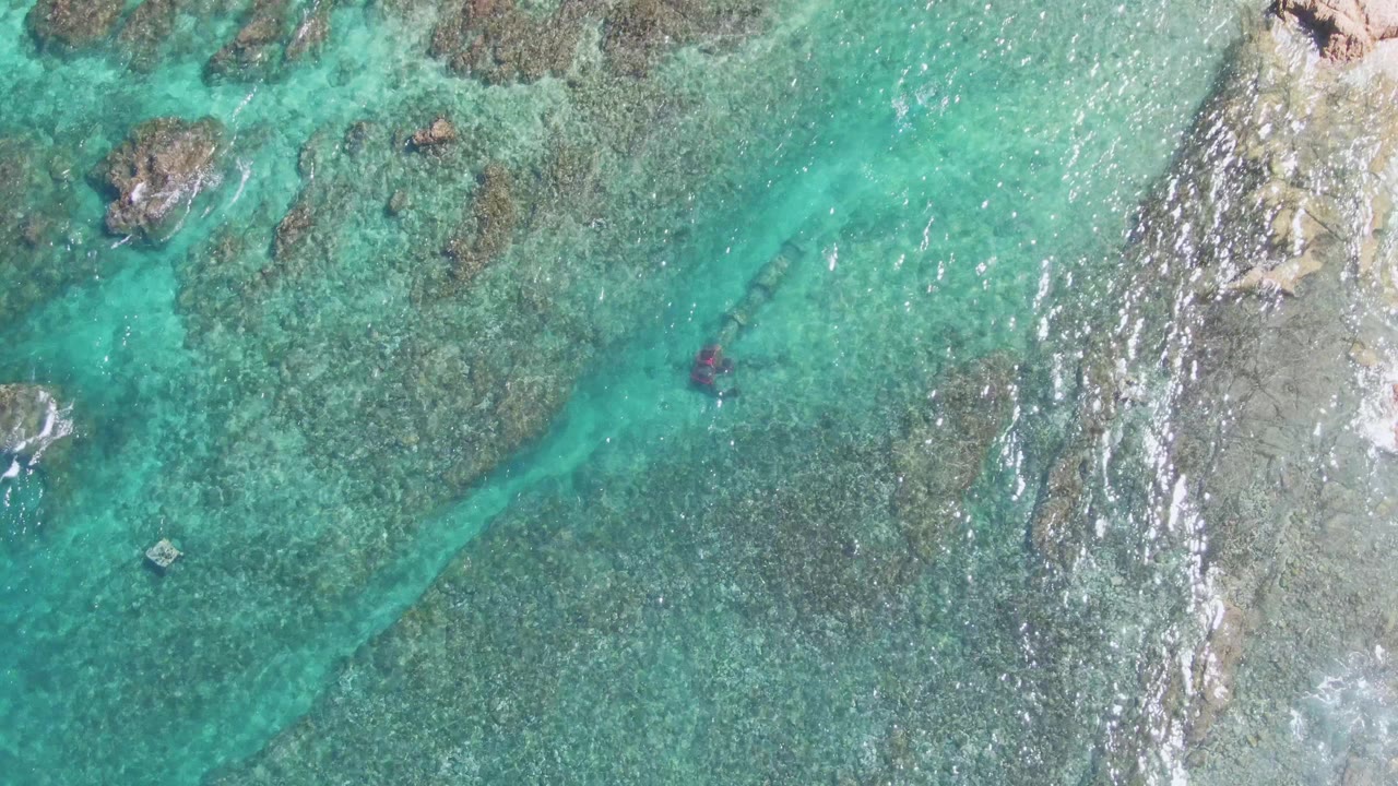 El Tule Beach Los Cabos shallows seen from above, with the swimmer form slightly farther through the clear water, reef texture below, and bright ripples along the right side.