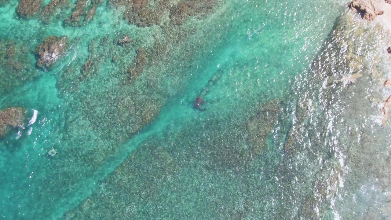 Top-down frame from El Tule Beach Los Cabos with turquoise water, darker reef patches, a small swimmer mark in the shallows, and foamy light streaks near the right edge.