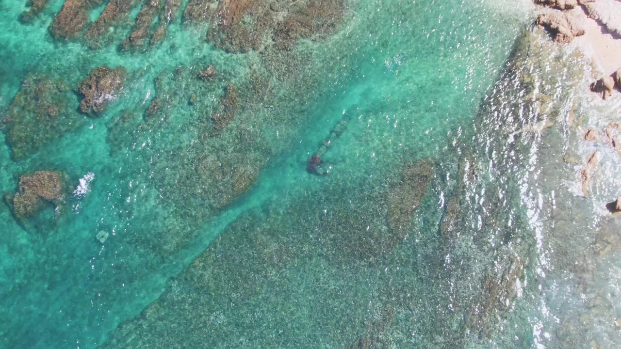 Rocky turquoise shallows with swimmer mark and shoreline rocks at the right edge.