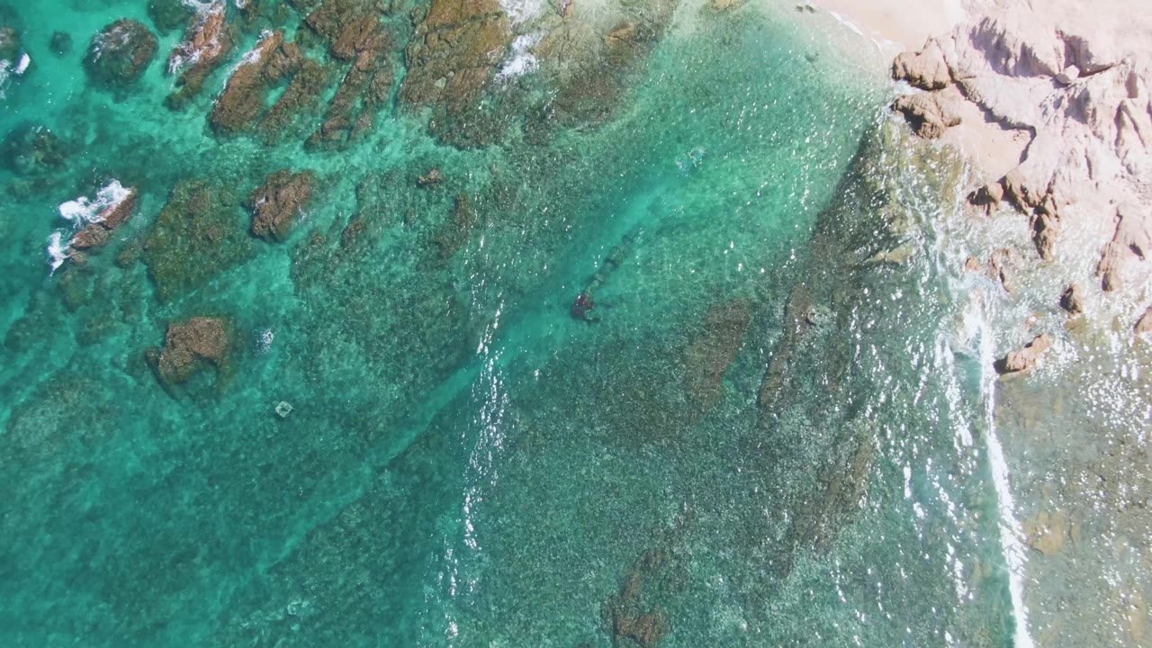 Clear turquoise shallows at El Tule Beach in Los Cabos ripple over darker submerged rocks, with the sandy rocky shore held along the right edge and no visible people.