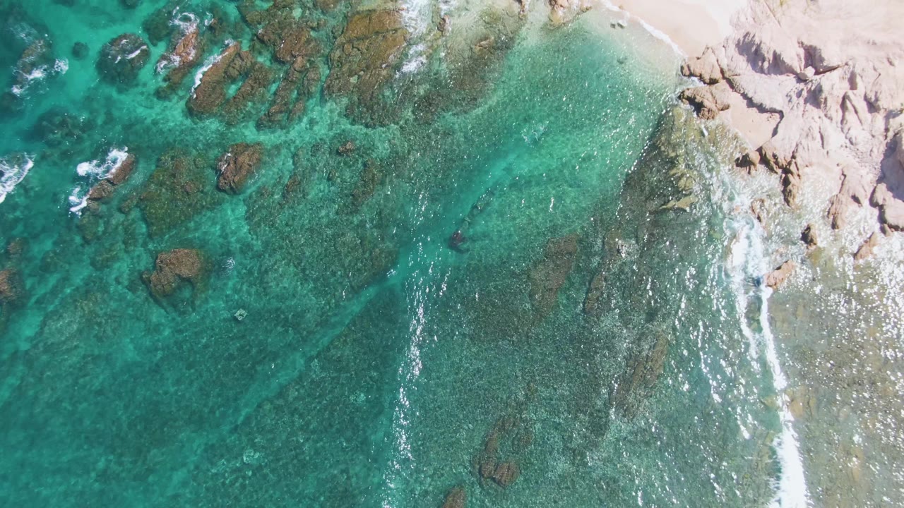 A top-down frame of El Tule Beach in Los Cabos shows a brighter foam streak along the right-side rocks, turquoise water across the center, and no visible people.