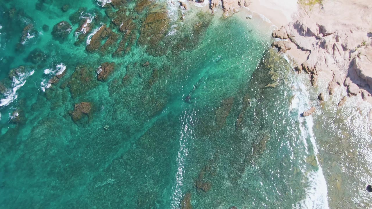 Drone-like overhead view at El Tule Beach in Los Cabos with clusters of dark rocks under turquoise surf, a pale rocky beach edge, and no visible people.