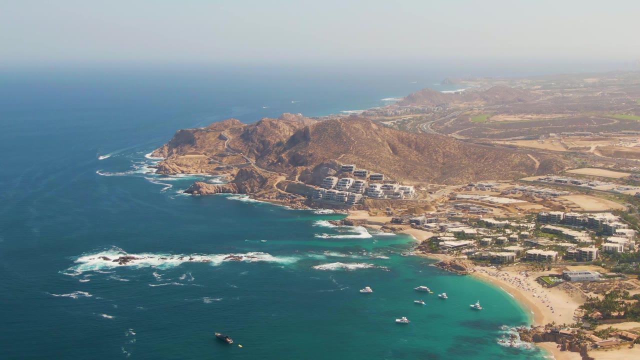 Opening aerial frame over El Tule Beach in Los Cabos, showing turquoise shallows, white surf, sandy shore, rocky headland, coastal buildings, and no visible people.