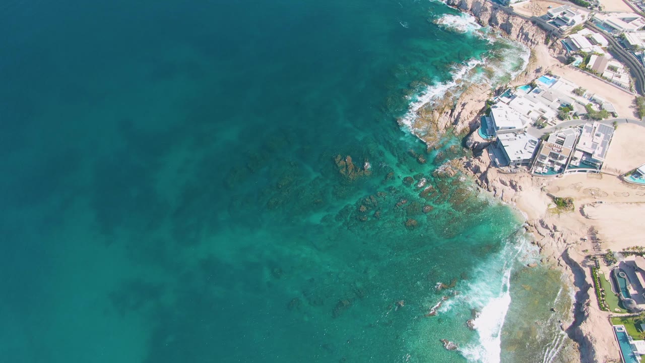 Aerial frame over El Tule Beach Los Cabos showing turquoise water on the left, white surf on rocks, sandy shore, and coastal buildings on the right with no visible people.