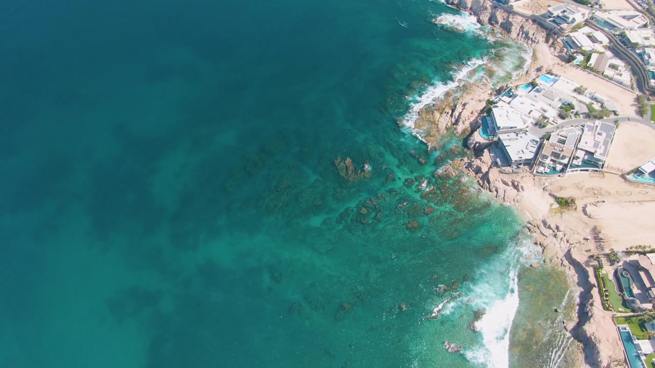 Aerial frame at El Tule Beach Los Cabos with clear blue-green water, a foam line breaking near the lower right rocks, and coastal buildings set back from the sand with no visible people.