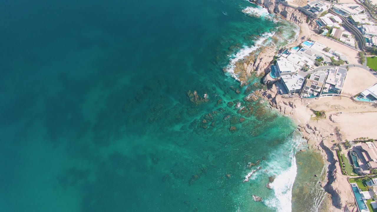 Overhead frame from El Tule Beach Los Cabos showing darker underwater rock patches, pale surf around the point, sandy beach, and clustered coastal buildings at the upper right with no visible people.