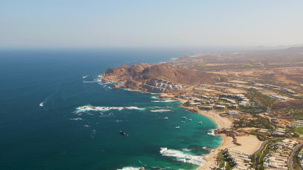Aerial view of turquoise bay, sandy beach, rocky hills, and coastal buildings.
