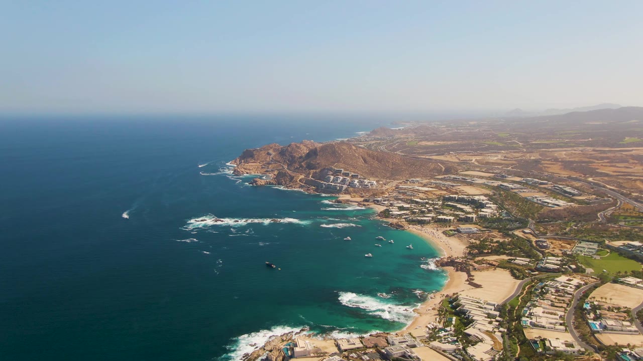 At 0:02, the aerial view over El Tule Beach holds the deep blue ocean on the left, turquoise shallows near the beach, coastal buildings to the right, rocky hills beyond, and no visible people.