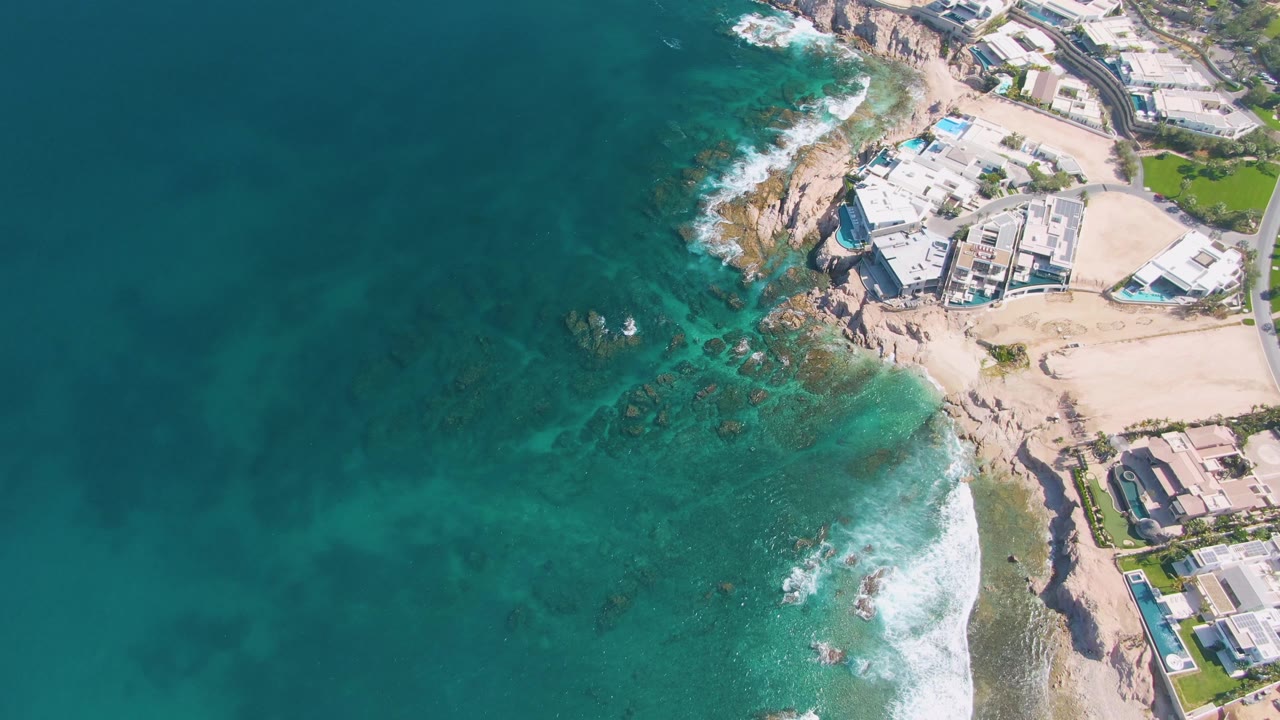Aerial frame over El Tule Beach in Los Cabos showing turquoise water, a sandy shore, rocky outcrops, beachfront buildings, pools, and no visible people.