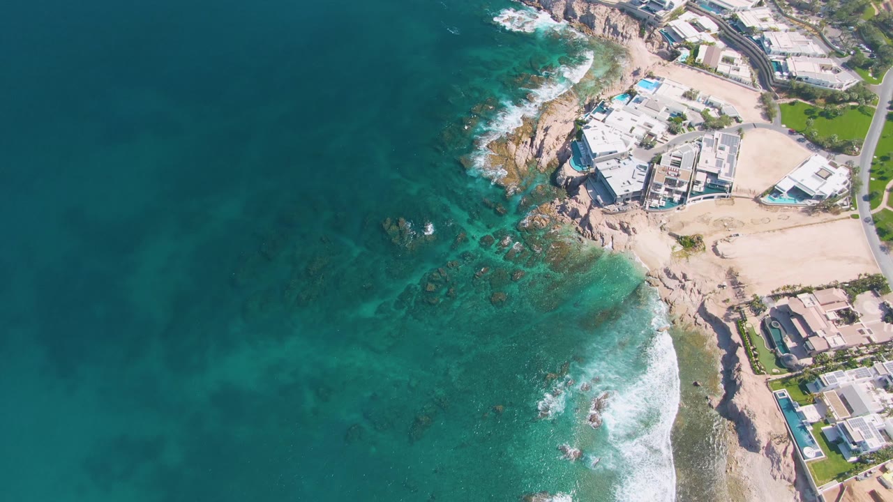 Drone view of waves breaking near rocks below beachside buildings and pools.