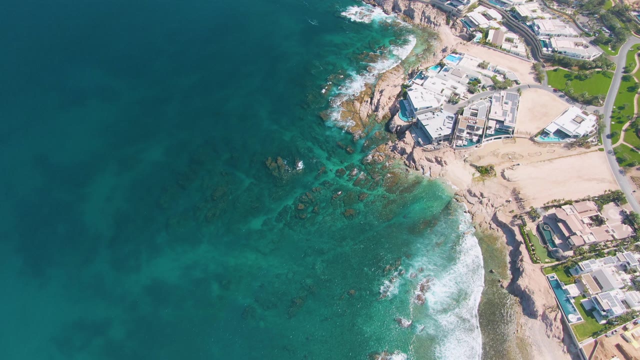 High drone frame over El Tule Beach in Los Cabos showing more open water on the left, surf over rocks near center, beachfront buildings and a road on the right, with no visible people.