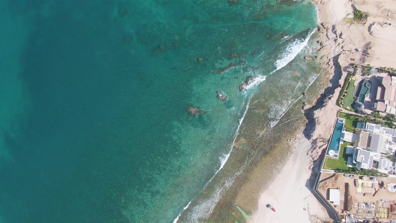 At 0:00, an aerial view over El Tule Beach in Los Cabos shows turquoise water on the left, surf crossing the sand, rocks near shore, and coastal buildings at the right edge.