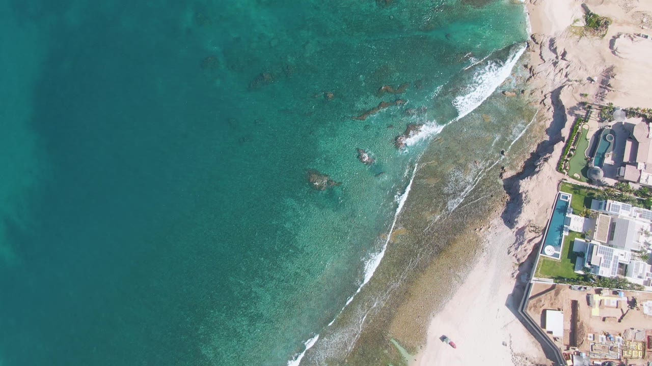 Aerial shoreline with turquoise ocean, white breaking waves, sand, rocks, and beach buildings.