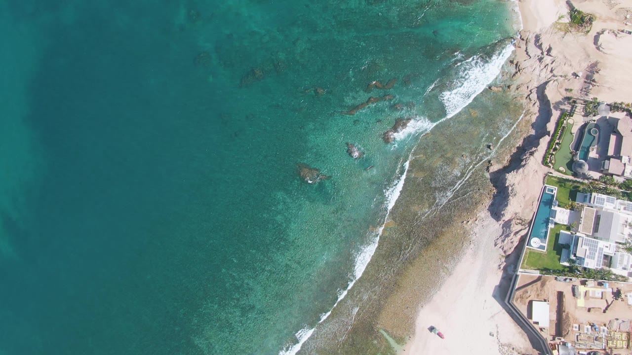 At 0:02, the aerial frame over El Tule Beach shows more white foam curling over the shallow reef while the sandy beach and coastal buildings stay along the right side.