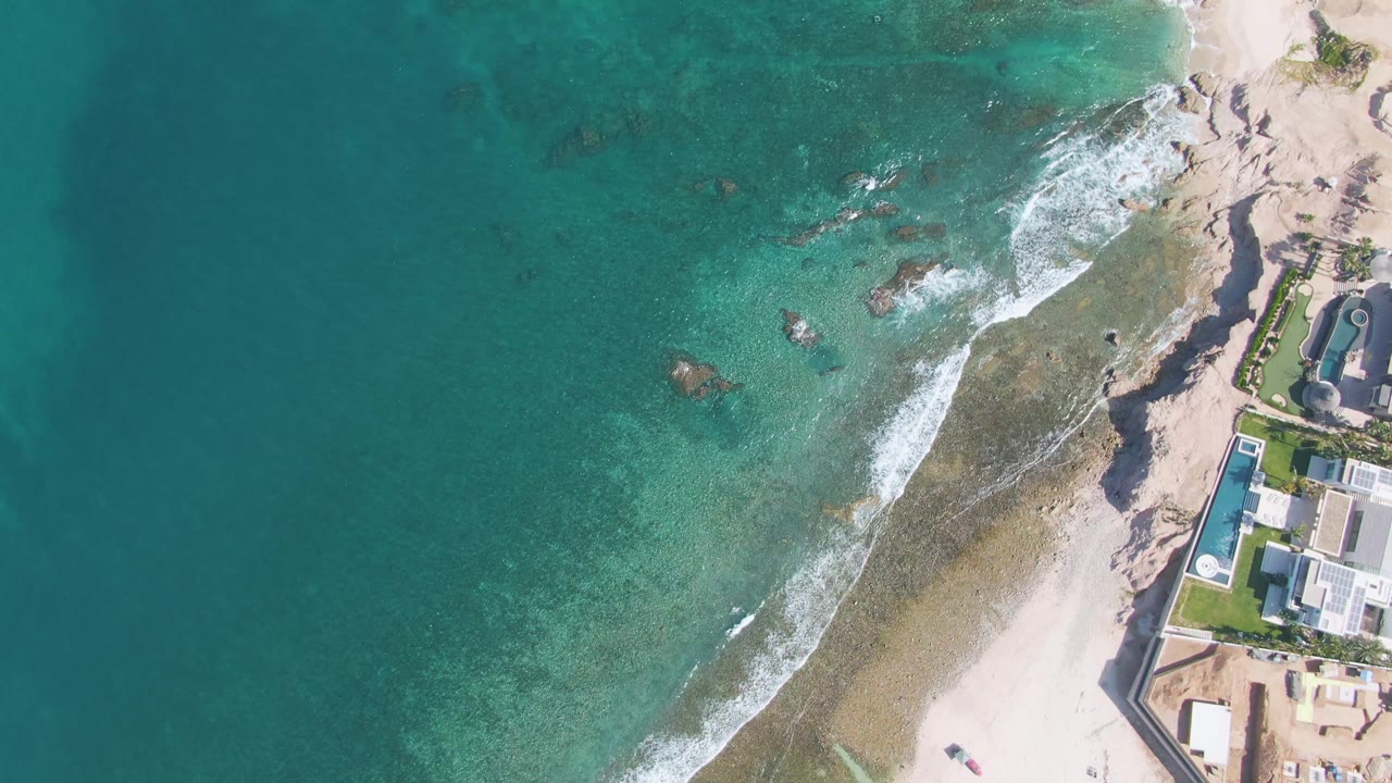 Aerial frame from El Tule Beach Los Cabos showing turquoise water, thin white surf, exposed rocks, pale sand, and coastal buildings along the right edge, with no visible people.