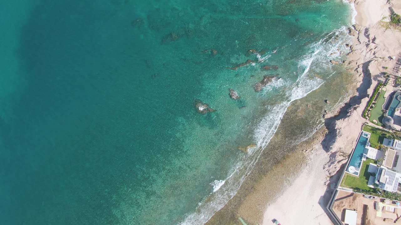 At 0:02, the aerial view over El Tule Beach Los Cabos holds on turquoise water and reef-like rocks while white foam gathers near the sandy shore and buildings remain cropped along the right edge, with no visible people.