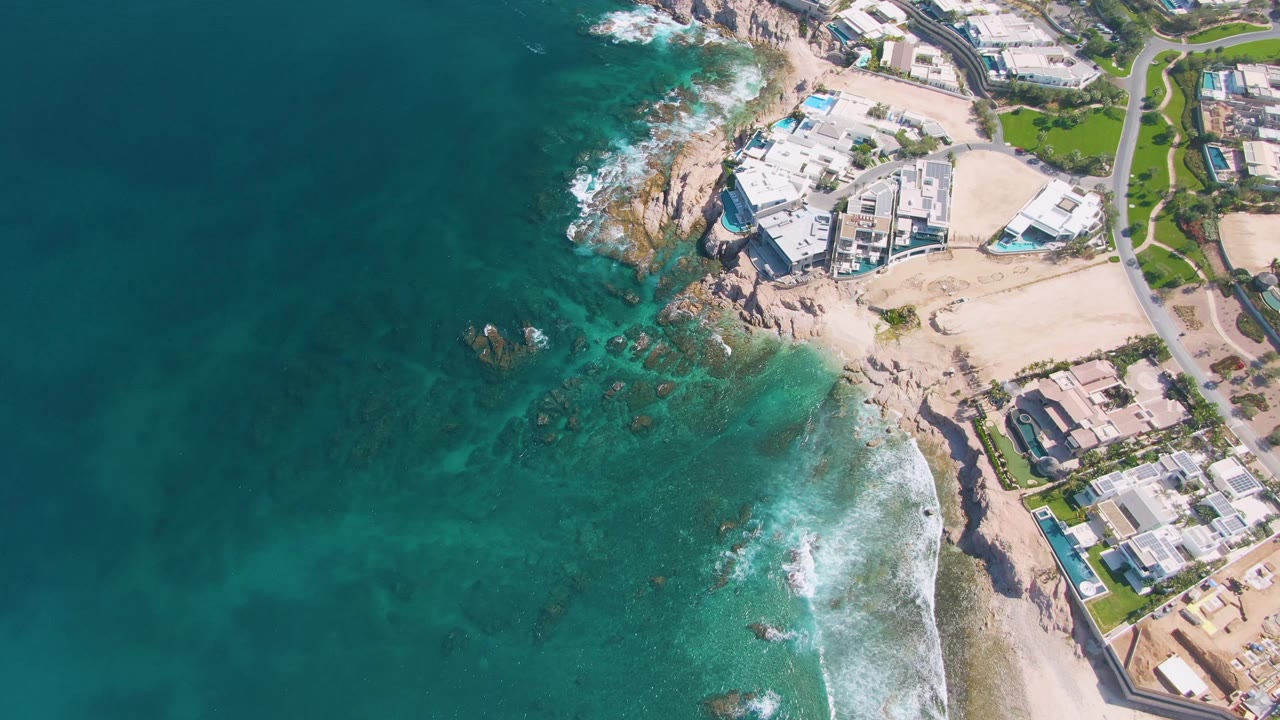 Aerial view over El Tule Beach in Los Cabos with turquoise water at left, white surf on rocks and sand, coastal buildings and road at right, and no visible people.