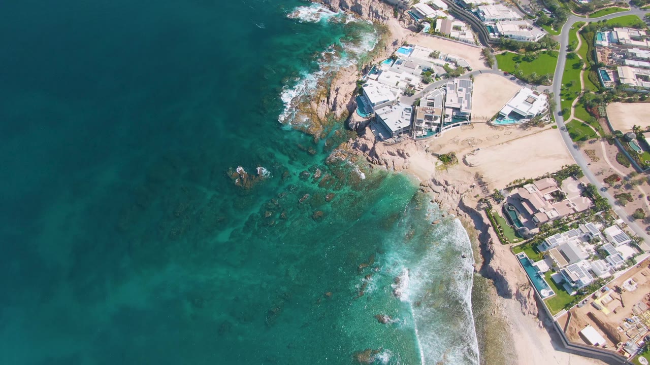Drone frame over El Tule Beach with darker ocean water on the left, clearer green water near the rocks, surf curling along the sandy edge, and no visible people.