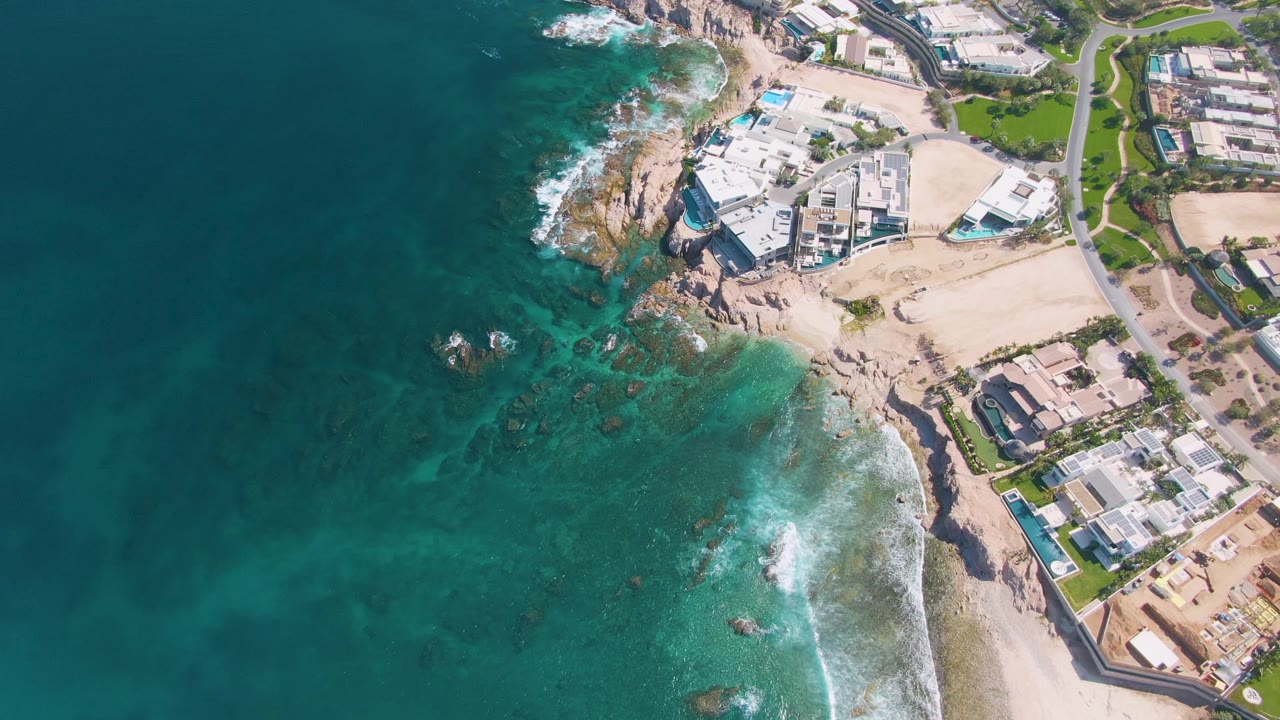 Aerial frame from El Tule Beach showing foamy waves around offshore rocks, pale sand, clustered coastal buildings, a road, green lawns, and no visible people.