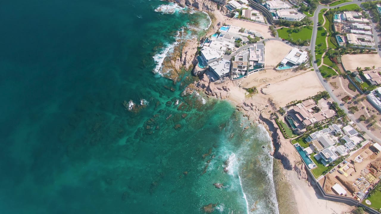 Wide overhead coastline with dark ocean, turquoise shallows, rocky point, and beach development.