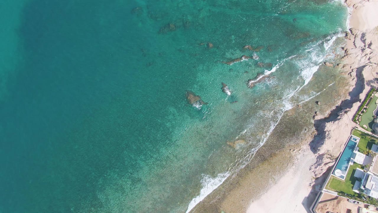 Aerial frame over El Tule Beach in Los Cabos showing turquoise water over submerged rocks, white foam near the sand, a poolside shore building at right, and no visible people.