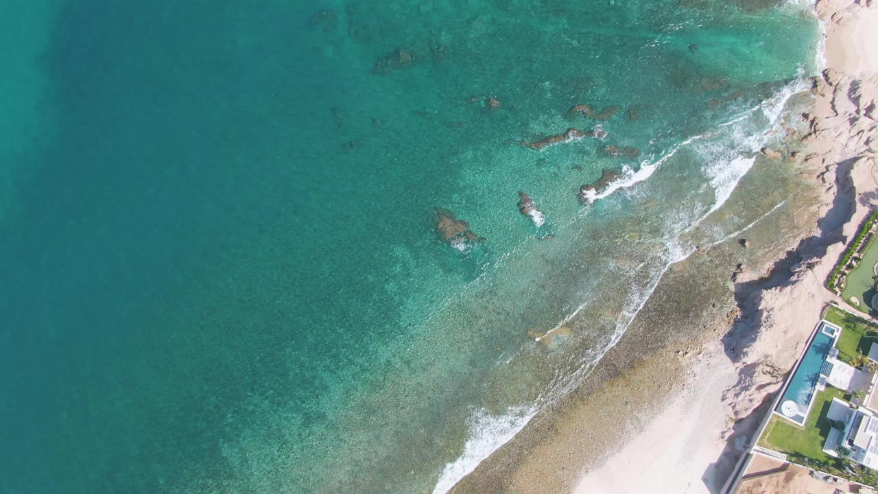 Aerial view at El Tule Beach in Los Cabos with clear turquoise water spreading across shallow rocks, surf tracing the beach, a pool and building on the right edge, and no visible people.