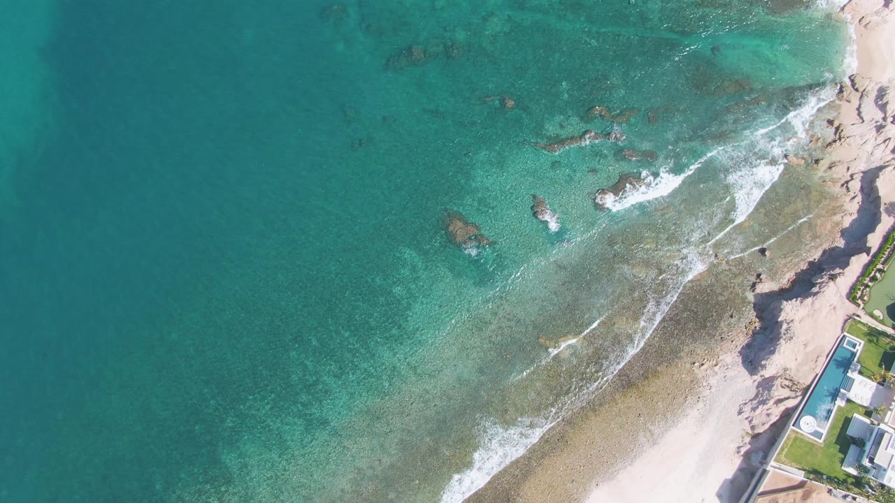 Top-down aerial frame from El Tule Beach in Los Cabos showing open turquoise water, darker rocks beneath the surface, foam breaking toward pale sand, a shore building at right, and no visible people.