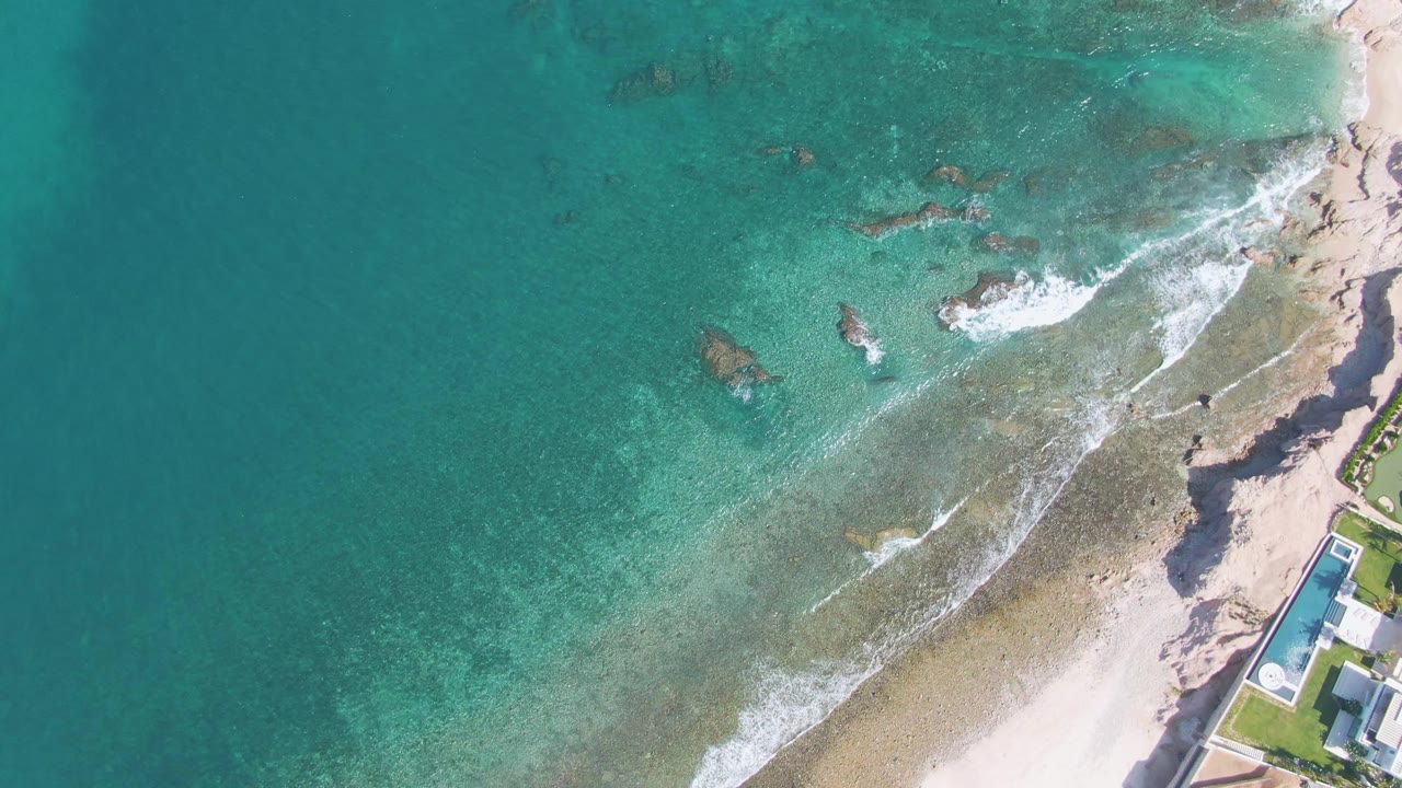 Aerial coastline with turquoise shallows, breaking waves, rocks, sand, and poolside property.