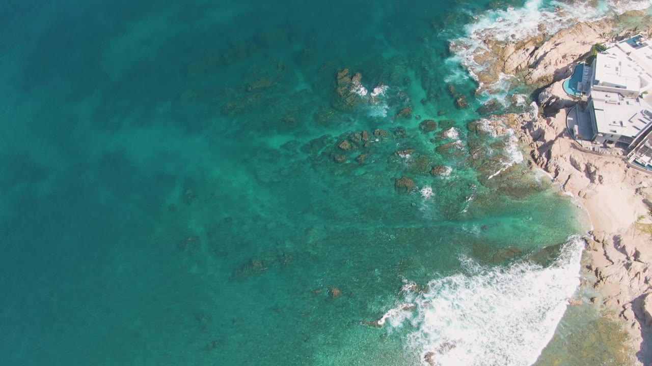 At El Tule Beach in Los Cabos, an overhead view shows turquoise water, submerged rocks, white surf on the lower shore, and a coastal building at the upper right with no visible people.