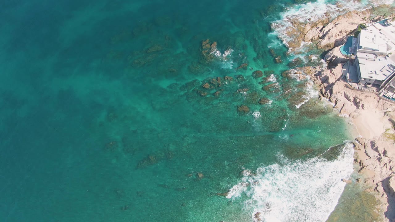At El Tule Beach in Los Cabos, white foam spreads farther across the lower rocks while clear blue-green water fills most of the frame and the coastal building remains at the upper right with no visible people.