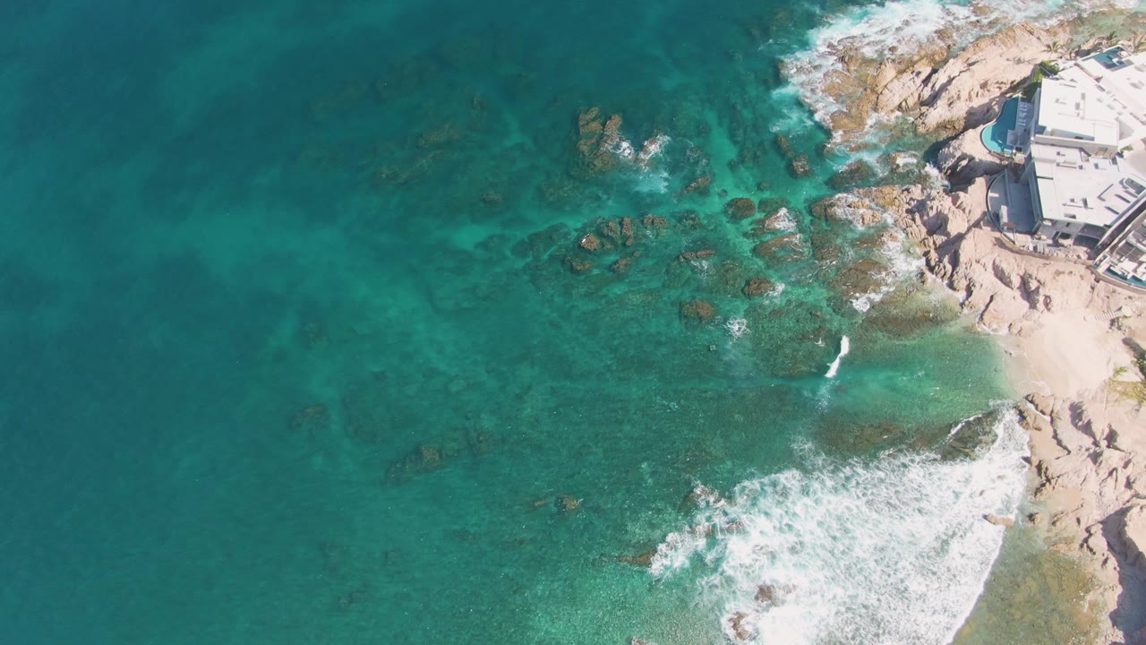 At El Tule Beach in Los Cabos, the surf line curls along the lower right beach edge, underwater rocks are visible through the water, and the coastal building sits partly cropped in the upper right with no visible people.