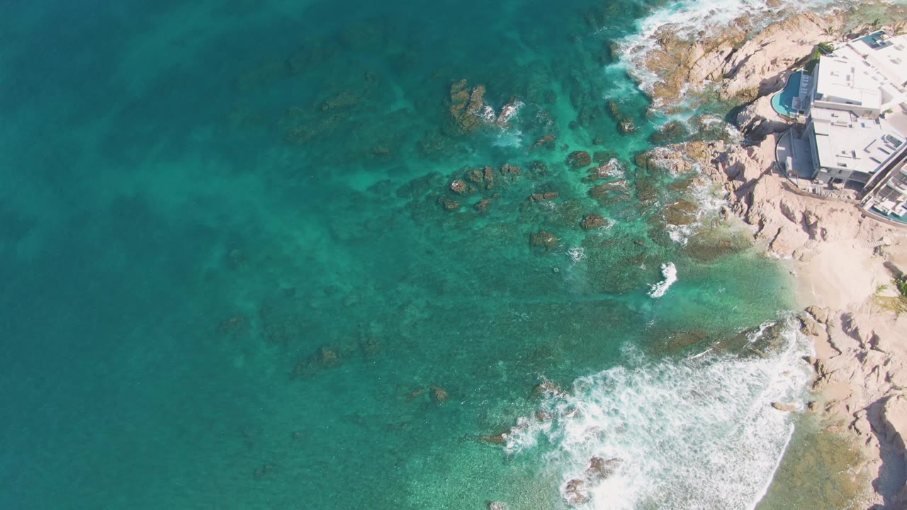 At El Tule Beach in Los Cabos, a compact white wave breaks near the sandy pocket on the right, with broad turquoise water and scattered rocks extending across the frame and no visible people.