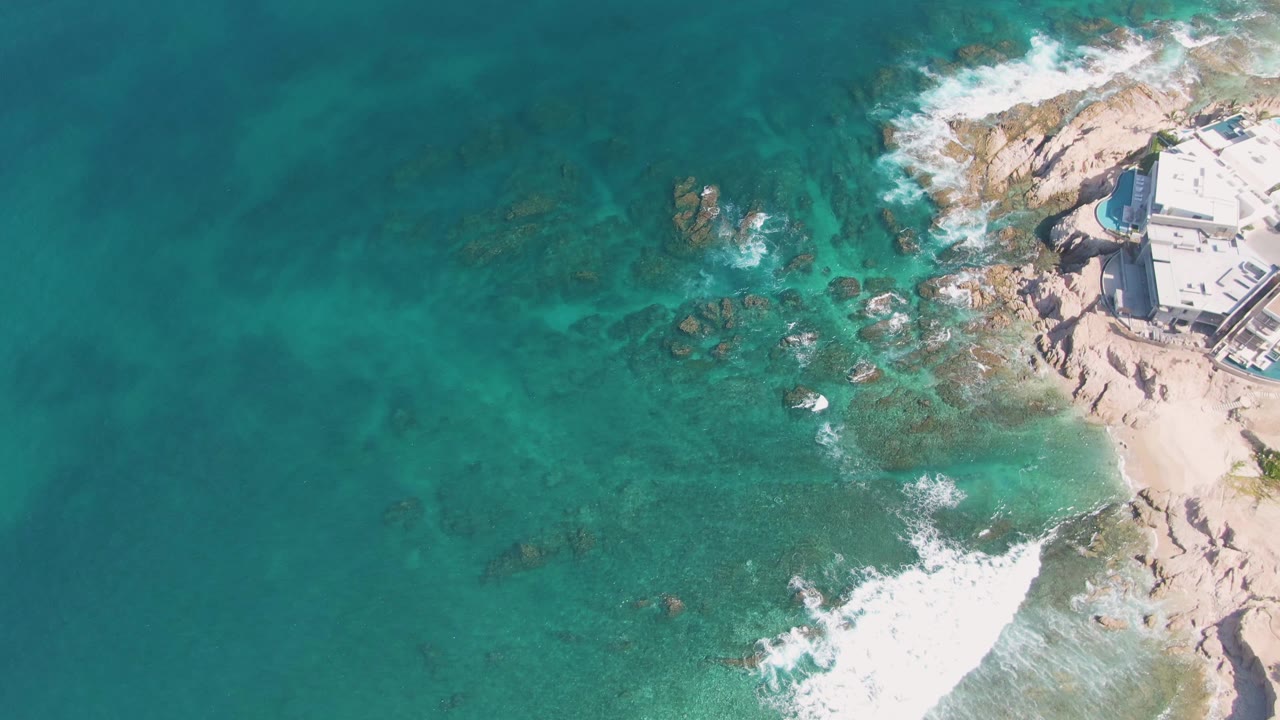 Overhead frame from El Tule Beach in Los Cabos showing turquoise water, submerged rocks, white surf foam, pale shoreline, partial coastal buildings, and no visible people.